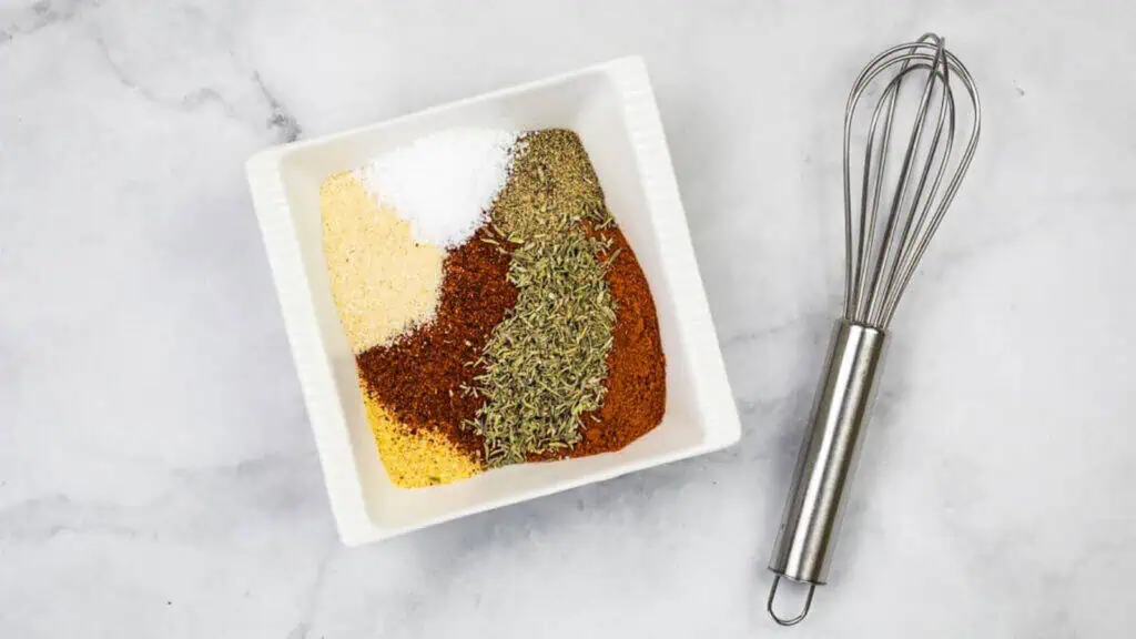 A square white bowl with six different spices arranged separately, next to a metal whisk on a light marble surface.