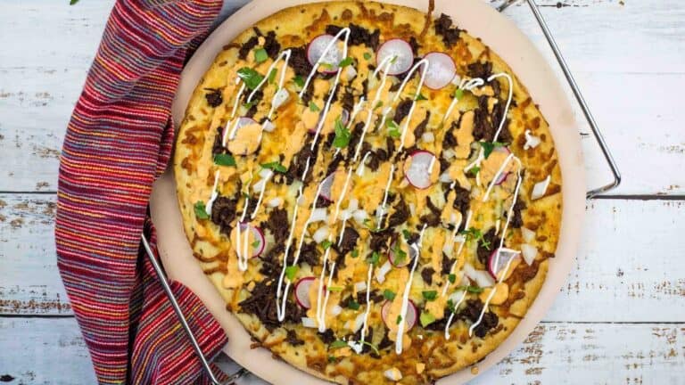 A cooked pizza topped with ground meat, cheese, sliced radishes, chopped herbs, and drizzled with white sauce on a round pizza stone next to a multicolored cloth.