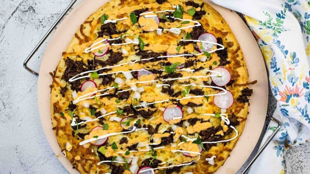 A cooked pizza topped with shredded meat, sliced radishes, chopped onions, herbs, cheese, and drizzled with a white sauce, served on a round tray next to a floral napkin.