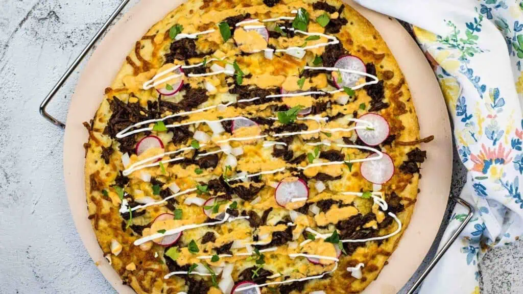A cooked pizza topped with shredded meat, sliced radishes, chopped onions, herbs, cheese, and drizzled with a white sauce, served on a round tray next to a floral napkin.