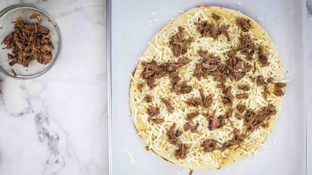 Uncooked pizza crust topped with shredded cheese and pulled meat sits on a baking sheet; a bowl of extra pulled meat is next to it on a marble surface.
