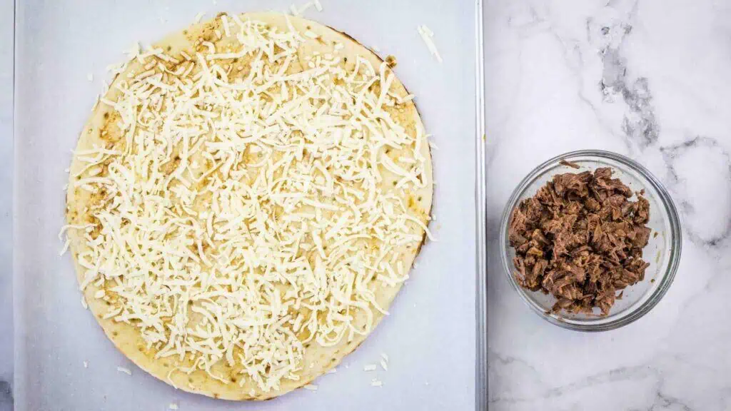 A pizza crust topped with shredded cheese sits on a baking sheet next to a bowl filled with cooked shredded meat on a marble countertop.