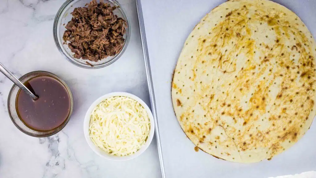 A baking sheet with a pizza crust, a bowl of shredded cheese, a bowl of shredded meat, and a bowl of sauce with a spoon on a marble surface.