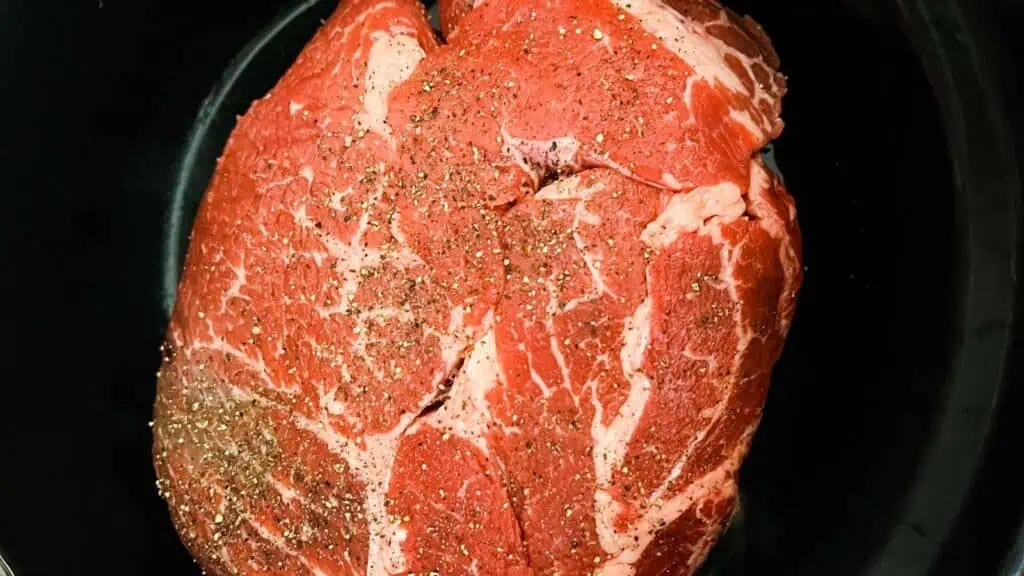 A raw, marbled beef roast with pepper and seasoning sits in a black slow cooker.