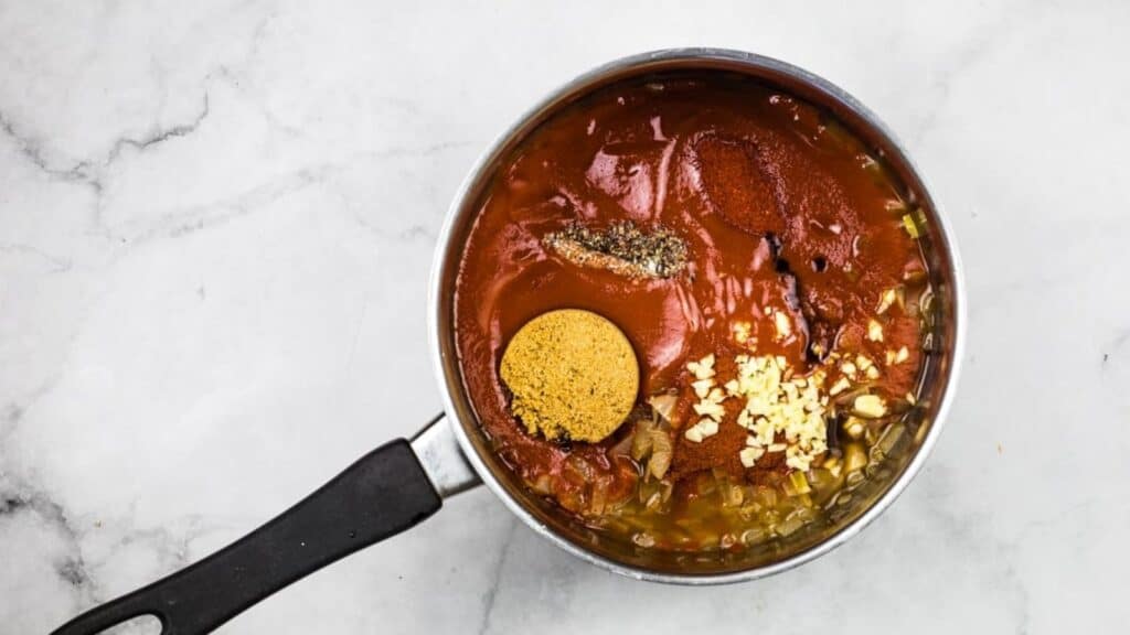 A saucepan on a marble surface filled with tomato sauce, brown sugar, minced garlic, herbs, and spices before mixing.