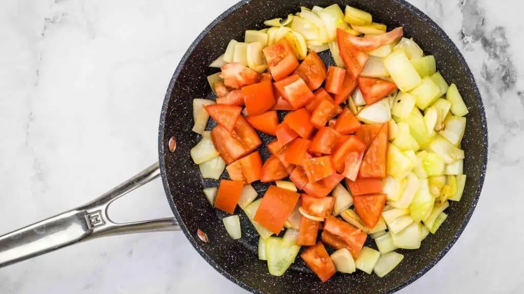 A frying pan containing chopped onions, celery, and tomatoes on a light-colored surface.