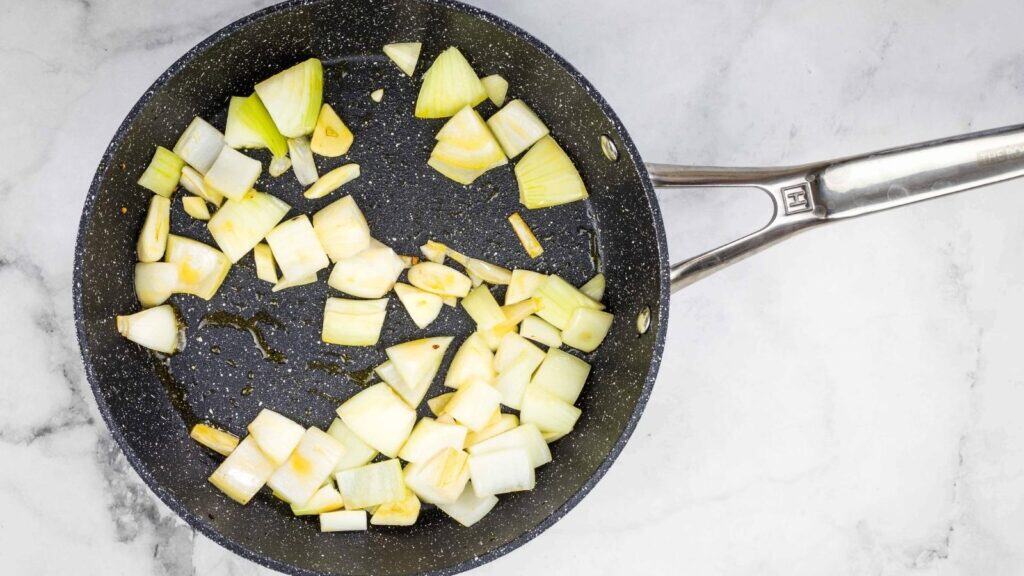 Diced onions and garlic cooking in a black nonstick frying pan with some oil on a marble surface.
