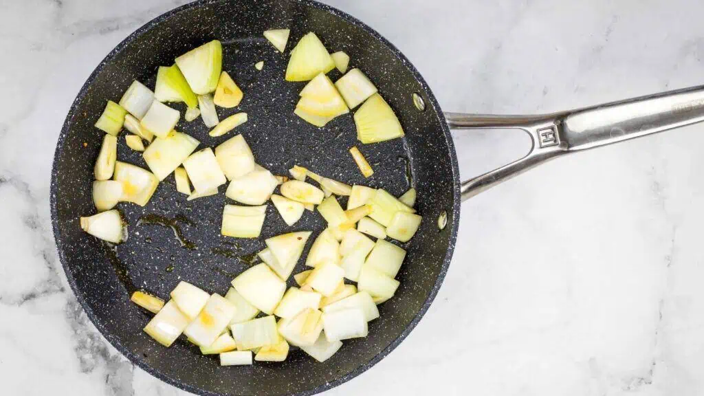 Diced onions and garlic cooking in a black nonstick frying pan with some oil on a marble surface.
