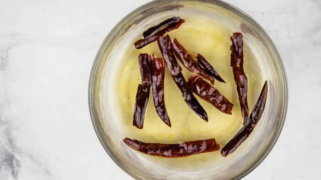 A glass bowl containing melted butter with several whole dried red chilies floating on top, viewed from above on a light surface.