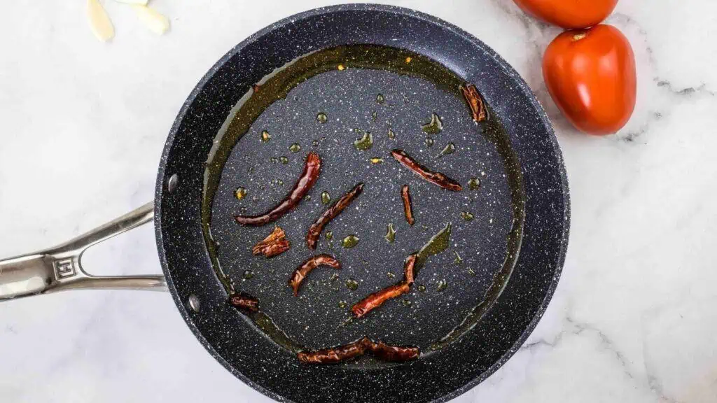 A frying pan with oil and dried red chilies on a marble surface, with two tomatoes and garlic cloves nearby.