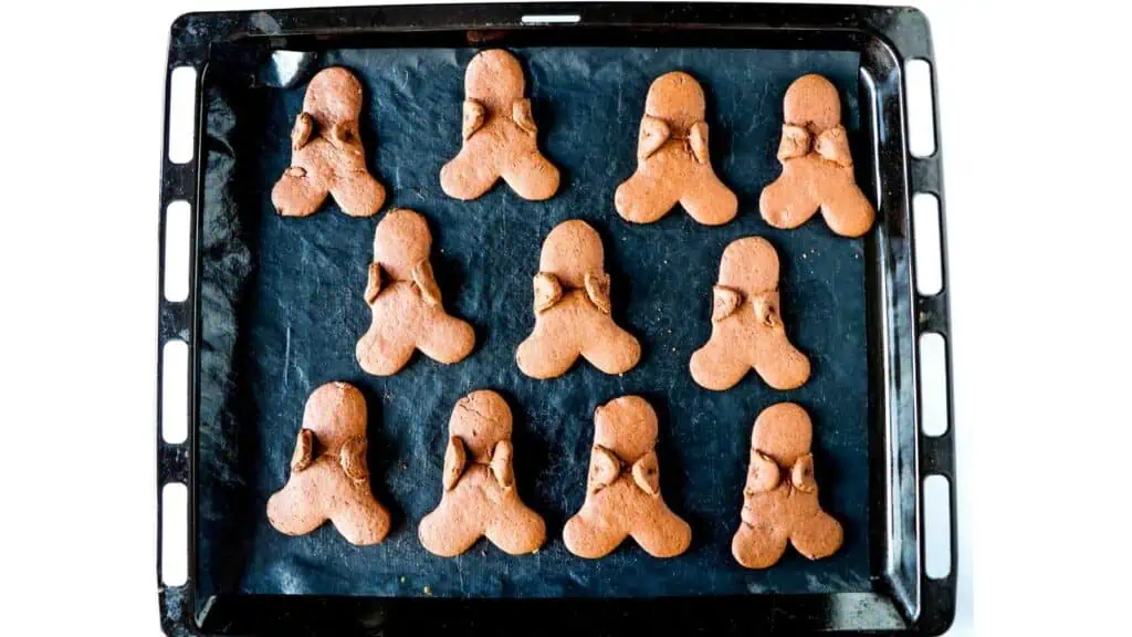 A baking tray with twelve brown cookies in an unusual shape, arranged in a grid on black parchment paper.