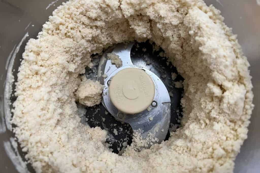 Food processor bowl with a metal blade attachment, containing a coarse, crumbly mixture of flour and fat, likely the beginning stage of making dough or pastry.