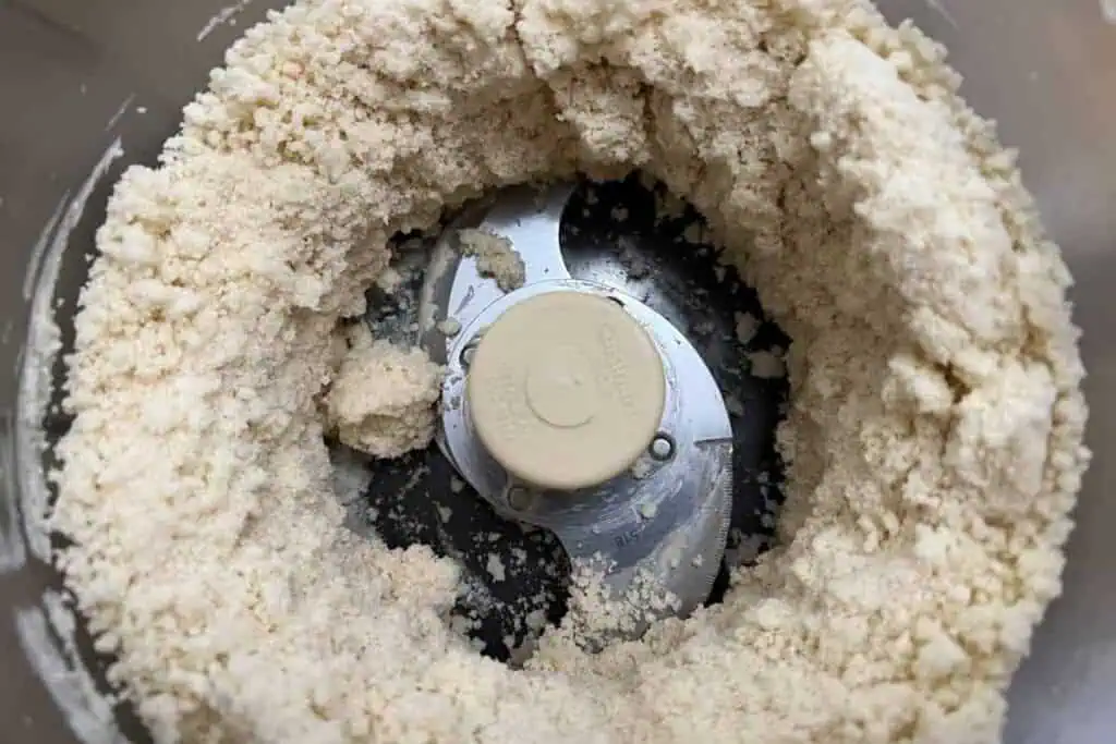 Food processor bowl with a metal blade attachment, containing a coarse, crumbly mixture of flour and fat, likely the beginning stage of making dough or pastry.