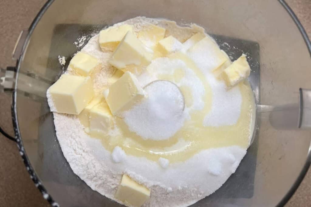Cubed butter, flour, and sugar in a food processor bowl, ready to be mixed for baking.