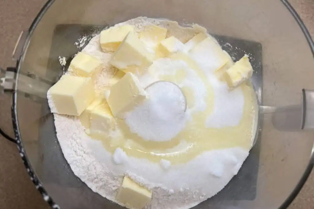 Cubed butter, flour, and sugar in a food processor bowl, ready to be mixed for baking.