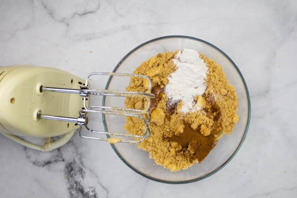 A glass bowl with brown sugar, cinnamon, and flour next to a hand mixer on a marble countertop.