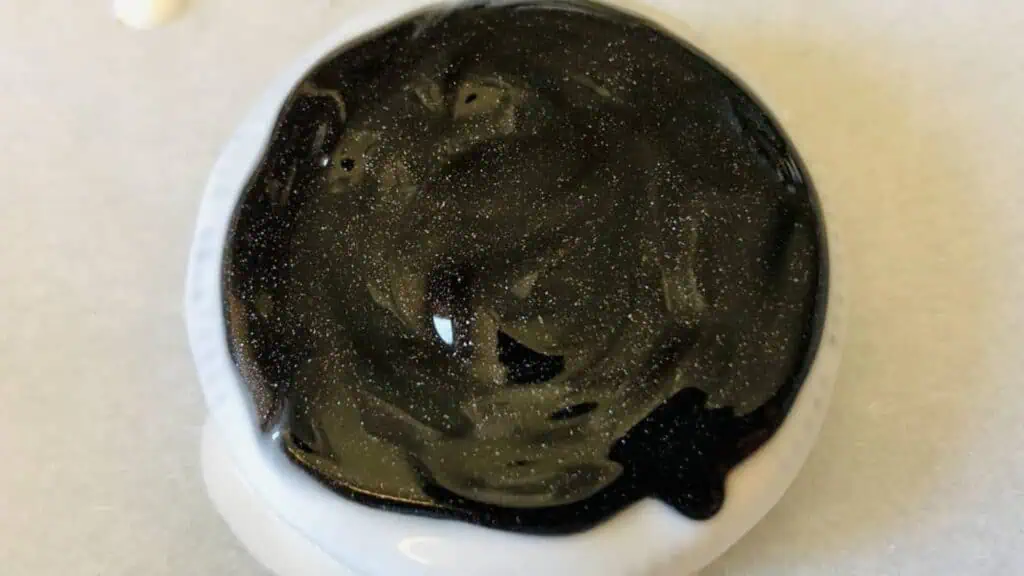 A round cookie with a glossy black icing on top, featuring subtle glitter-like sparkles, sits on a white surface.