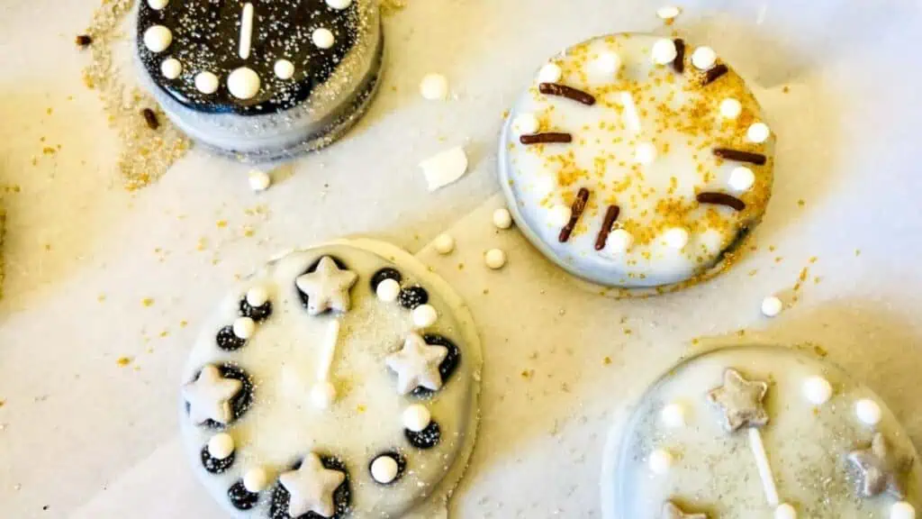 Iced cookies decorated like clock faces, featuring white, gold, and black icing with star and round sprinkles, arranged on a white surface.