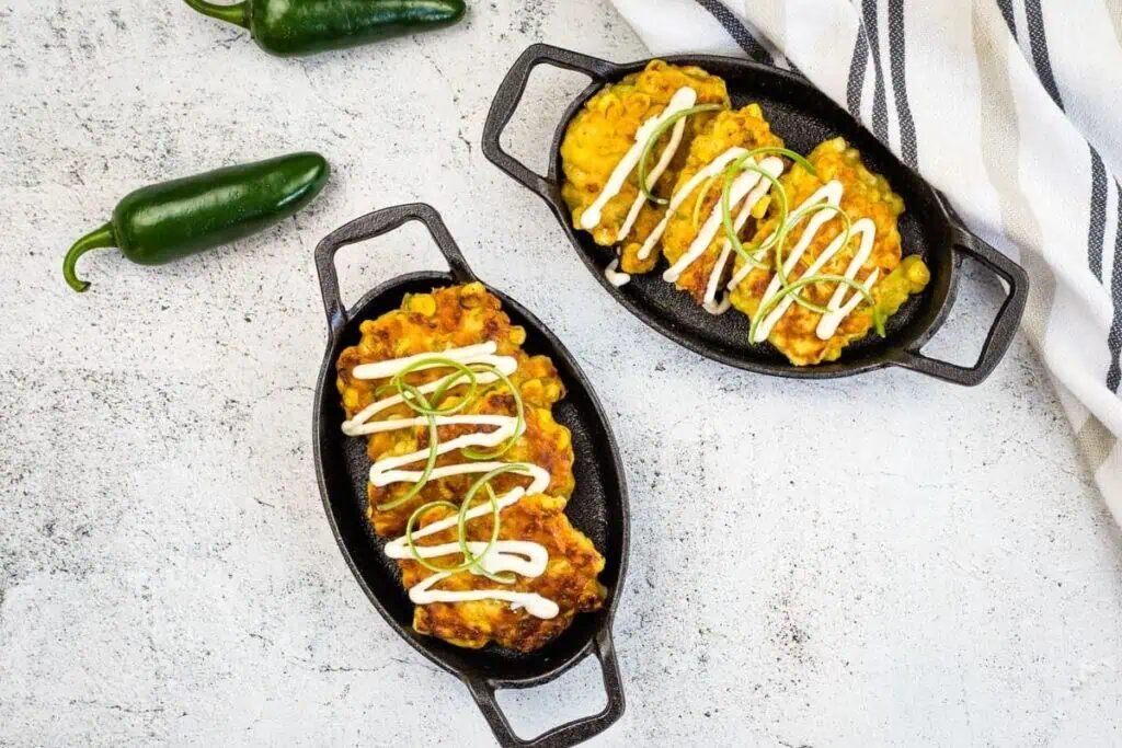 Two cast iron dishes with corn fritters topped with sour cream and green onions, next to two whole jalapeños and a white striped cloth on a light surface.