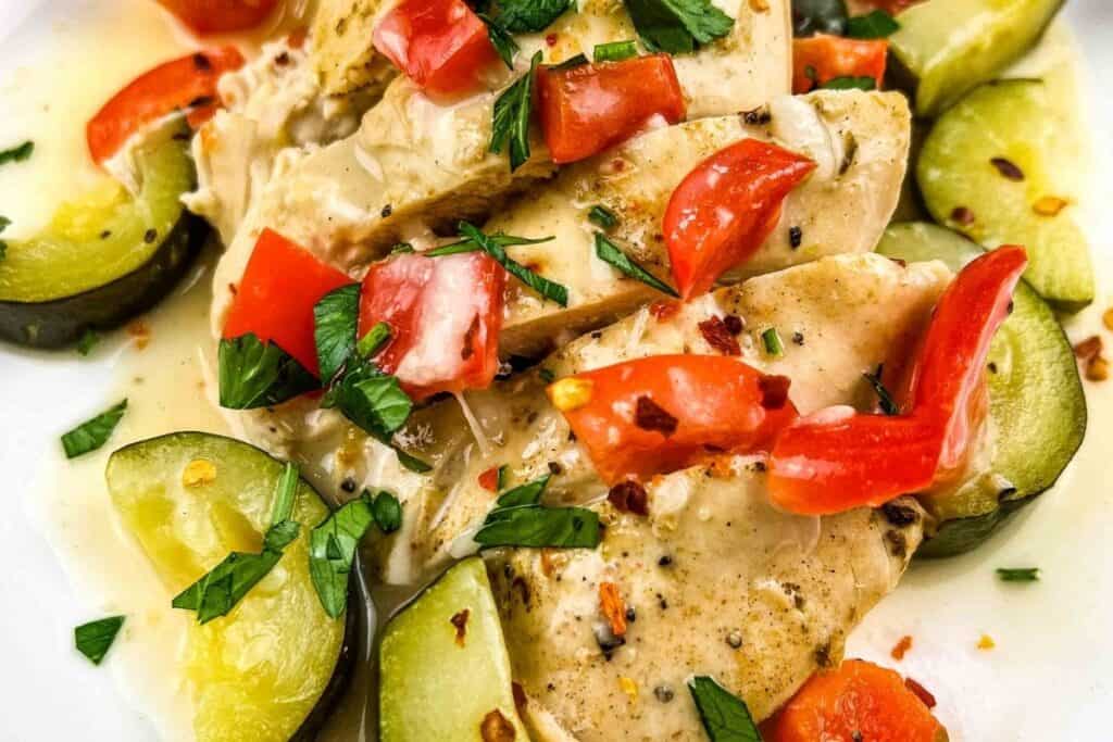 Sliced cooked chicken breast with zucchini, diced red bell pepper, and chopped parsley, served in a creamy sauce on a white plate.