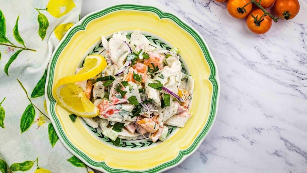 A bowl of creamy vegetable salad garnished with chopped herbs and lemon slices, placed on a marble surface next to cherry tomatoes and a lemon-patterned napkin.