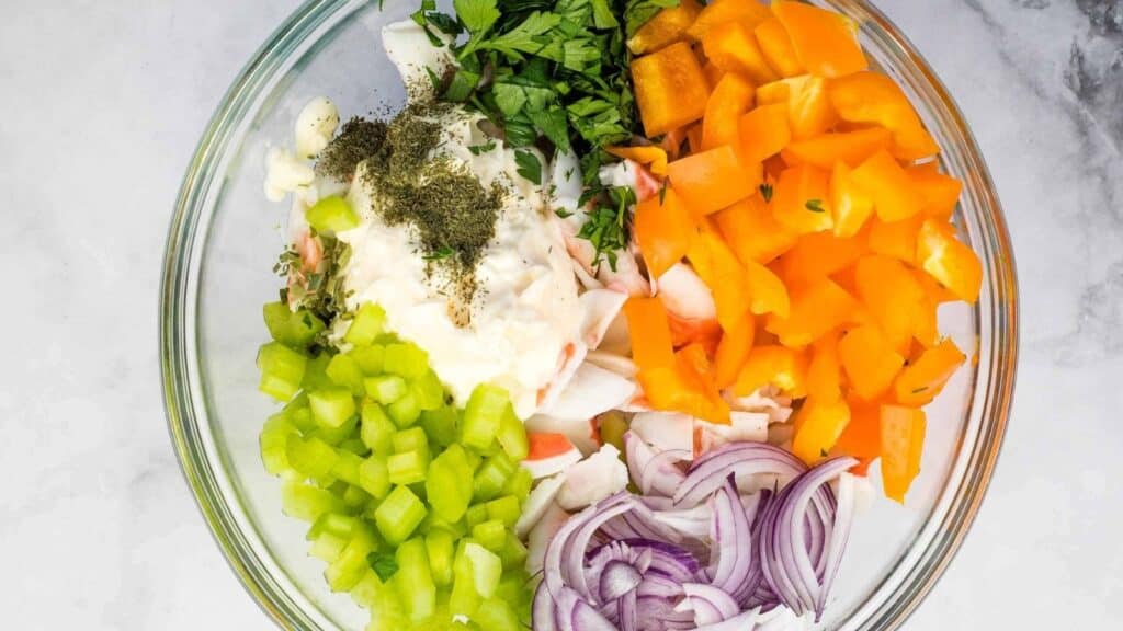 A glass bowl containing chopped celery, orange bell pepper, red onion, parsley, imitation crab, mayonnaise, and dried herbs on a light surface.