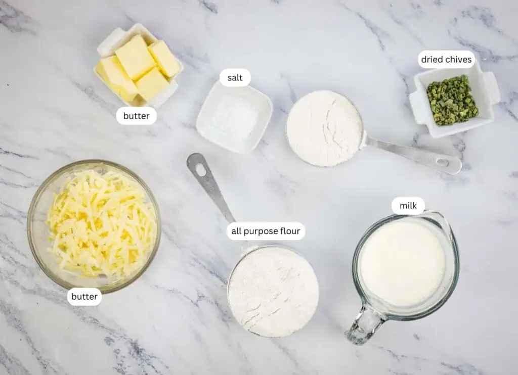 Ingredients on a marble counter: cubed butter, shredded butter, salt, dried chives, two bowls of all-purpose flour, and a measuring cup of milk.