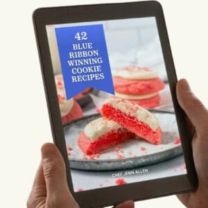 A person holds a tablet displaying an e-book cover titled "42 Blue Ribbon Winning Cookie Recipes" by Chef Jenn Allen, featuring pink and white cookies on a plate.