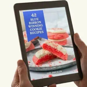 A person holds a tablet displaying an e-book cover titled "42 Blue Ribbon Winning Cookie Recipes" by Chef Jenn Allen, featuring pink and white cookies on a plate.