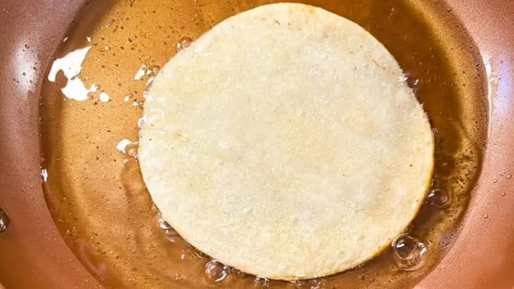 A corn tortilla is frying in hot oil in a copper-colored pan, surrounded by small bubbles.