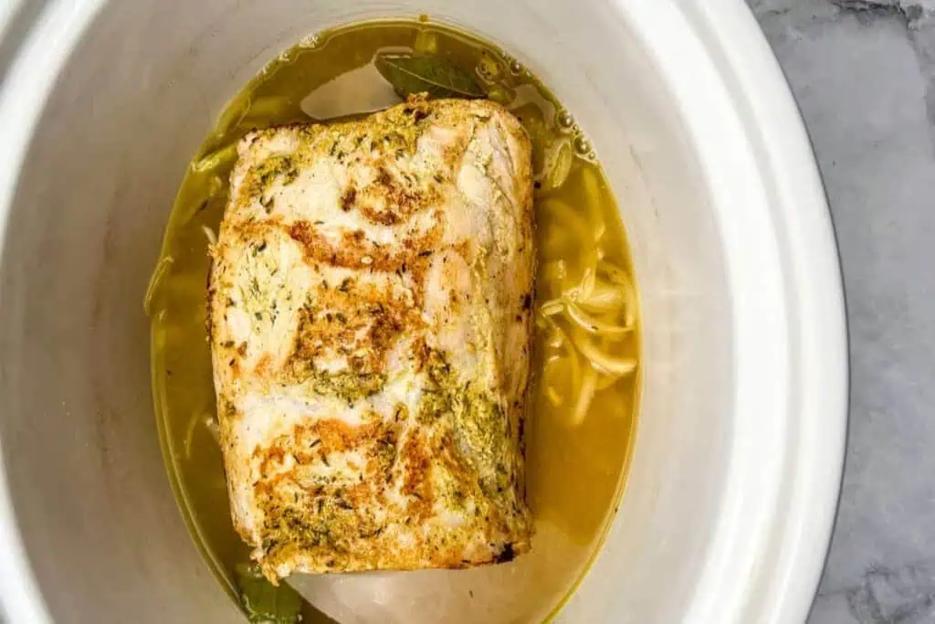 Cooked pork roast with seasoning in broth with bay leaves and onion slices inside a white slow cooker.