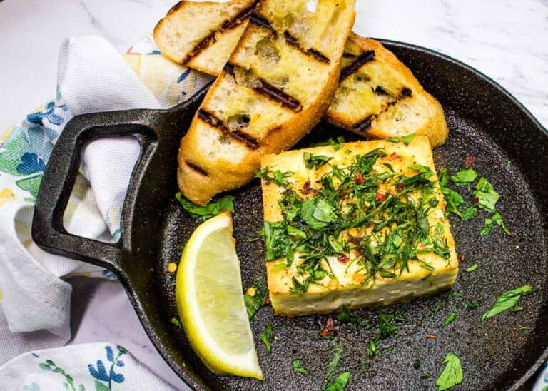 A slice of grilled bread and a herbed cheese block are served in a cast iron skillet with a lemon wedge and sprinkled parsley, alongside a white cloth with a floral pattern.