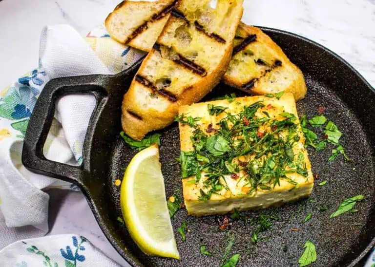A slice of grilled bread and a herbed cheese block are served in a cast iron skillet with a lemon wedge and sprinkled parsley, alongside a white cloth with a floral pattern.