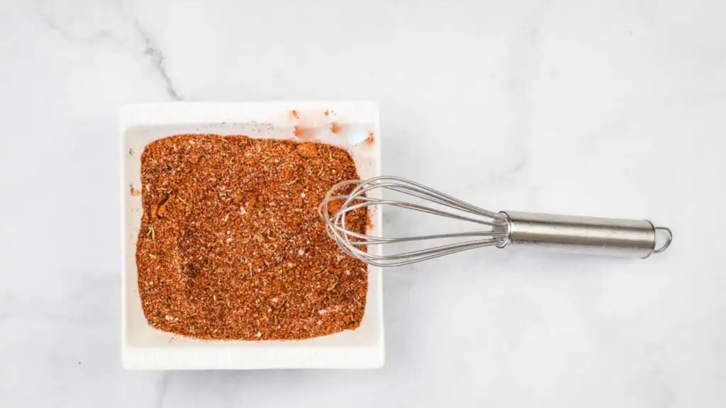 A metal whisk rests in a square white dish filled with a mixed dry seasoning blend on a white marble surface.