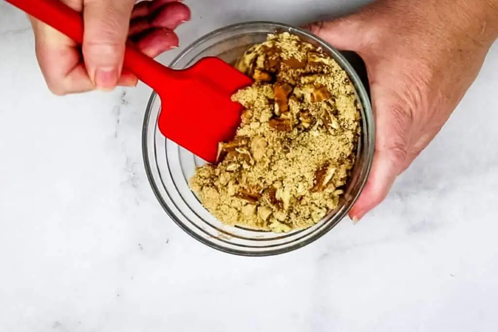 A person mixes brown sugar, cinnamon, and chopped pecans in a glass bowl with a red spatula.