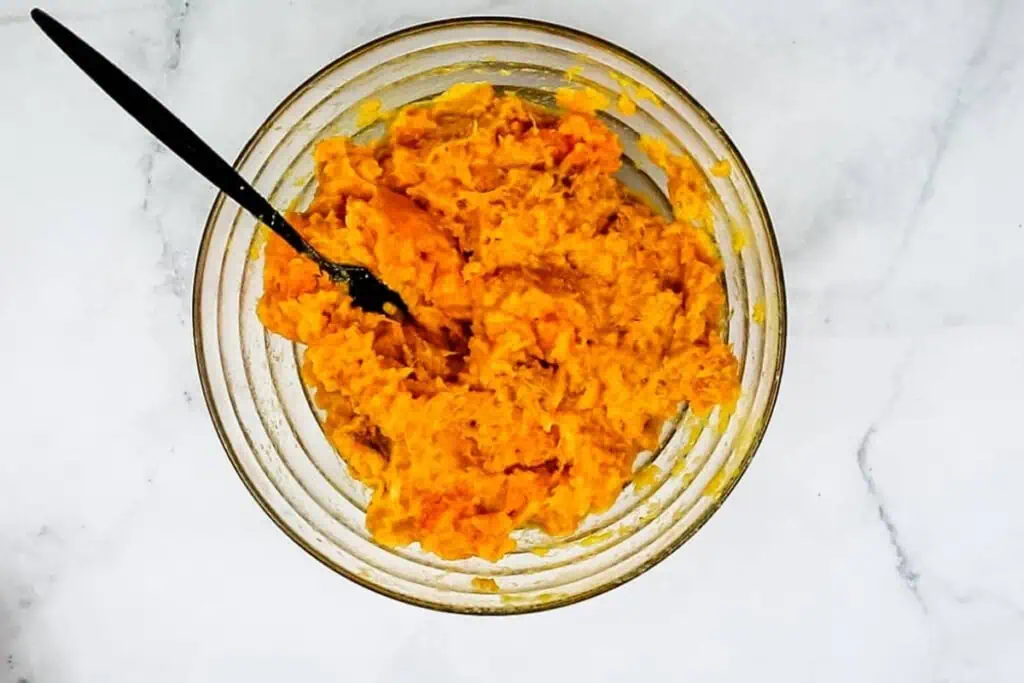 A glass bowl filled with mashed sweet potatoes and a black spoon, placed on a white marble surface.