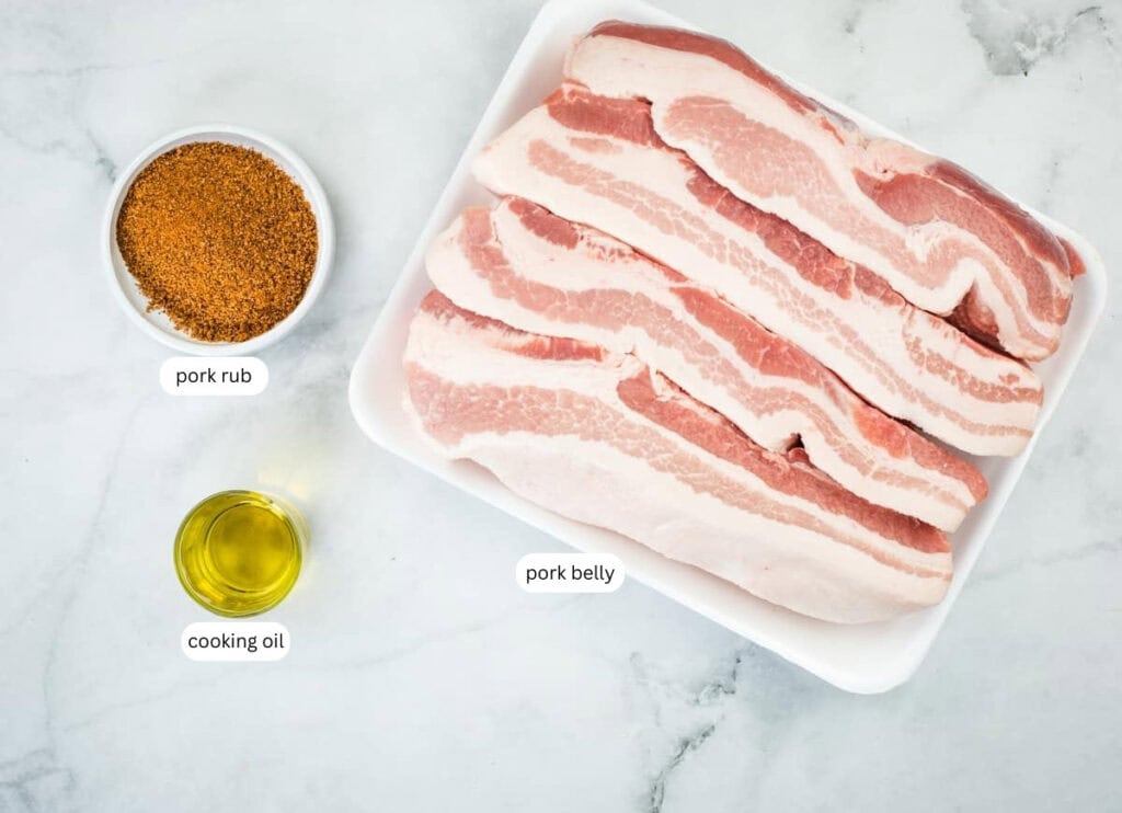 A tray of raw pork belly slices, a bowl of pork rub, and a small glass container of cooking oil on a white marble surface.