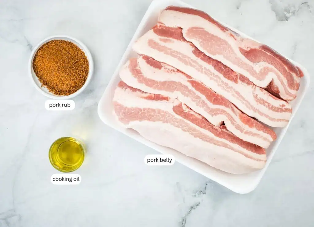 A tray of raw pork belly slices, a bowl of pork rub, and a small glass container of cooking oil on a white marble surface.