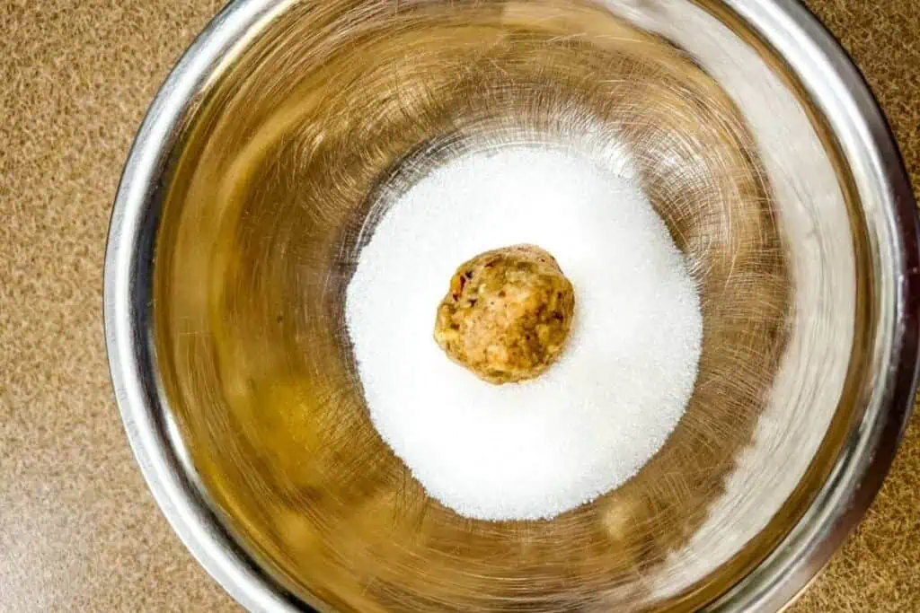 A metal bowl containing granulated white sugar with a single unbaked cookie dough ball placed on top.