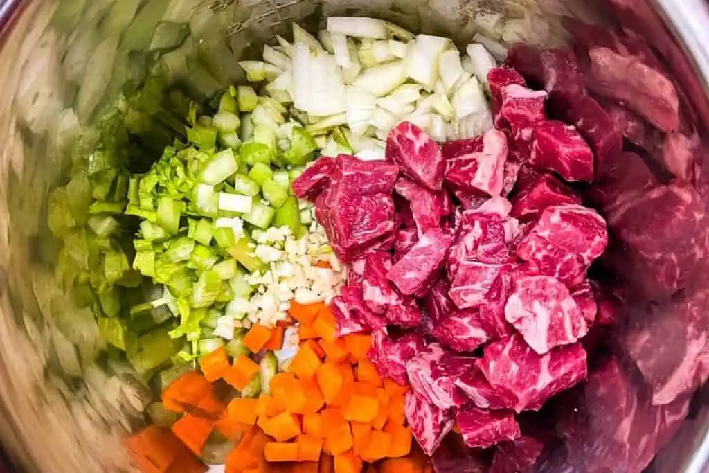 Chopped onions, celery, carrots, garlic, and raw beef chunks arranged in sections inside a metal mixing bowl.