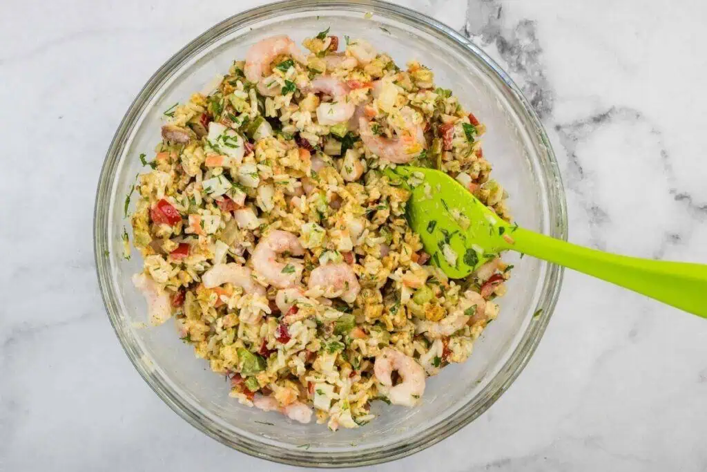 A glass bowl filled with shrimp and rice salad mixed with chopped vegetables and herbs, with a green spatula in the bowl.