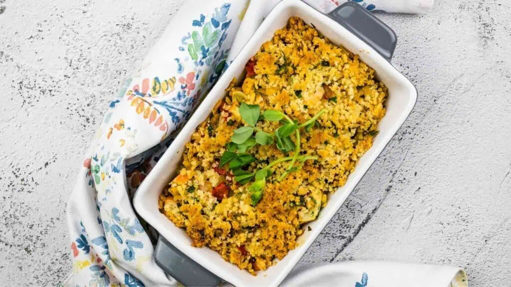 A rectangular baking dish filled with vegetable casserole topped with golden breadcrumbs, garnished with fresh greens, placed on a floral-patterned cloth.