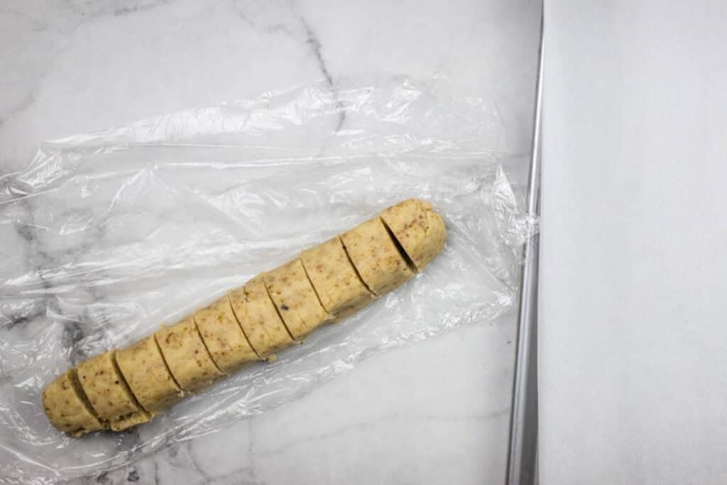 A log of cookie dough, partially sliced into round pieces, rests on plastic wrap on a marble surface next to a baking sheet lined with parchment paper.