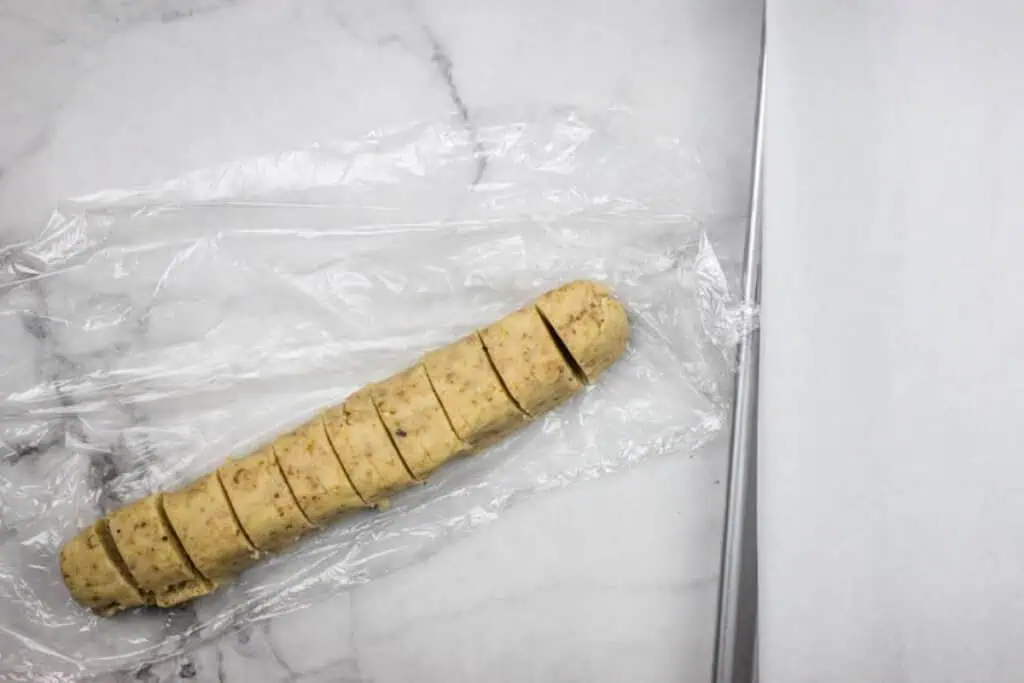 A log of cookie dough, partially sliced into round pieces, rests on plastic wrap on a marble surface next to a baking sheet lined with parchment paper.
