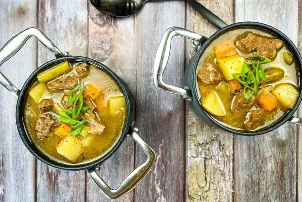 Two small black pots filled with beef stew containing chunks of beef, potatoes, carrots, and green onions, placed on a wooden surface with a metal spoon nearby.