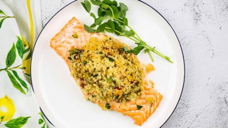 A fillet of salmon topped with a breadcrumb and herb mixture is served on a white plate with sprigs of greens, next to a patterned napkin.