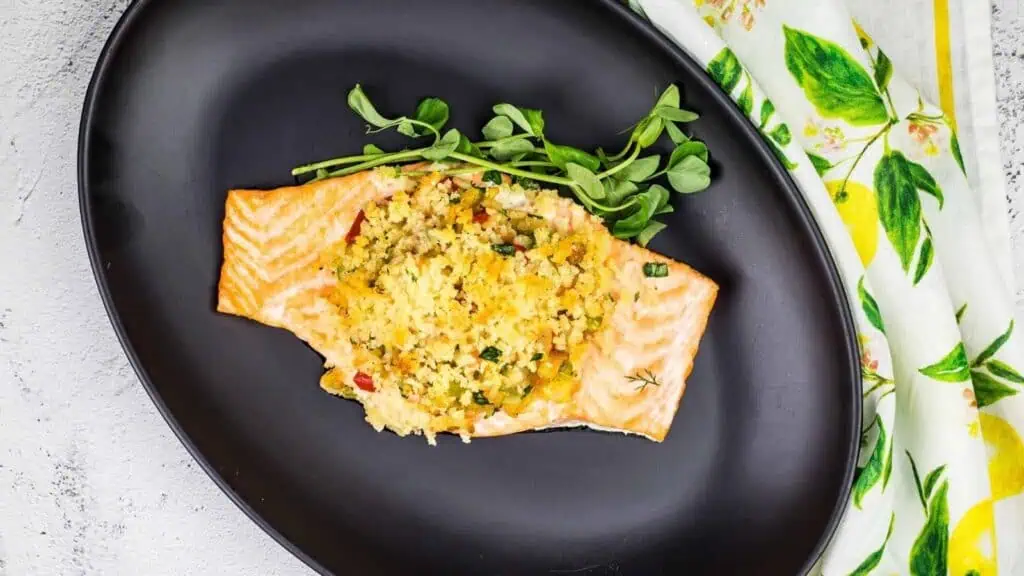 Baked salmon fillet topped with a breadcrumb and vegetable mixture, garnished with pea shoots, served on a black oval plate.