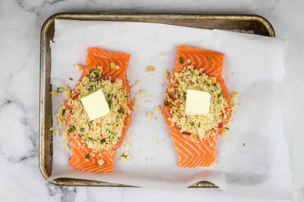 Two uncooked salmon fillets on a parchment-lined baking sheet, topped with a crumb mixture and a pat of butter each.