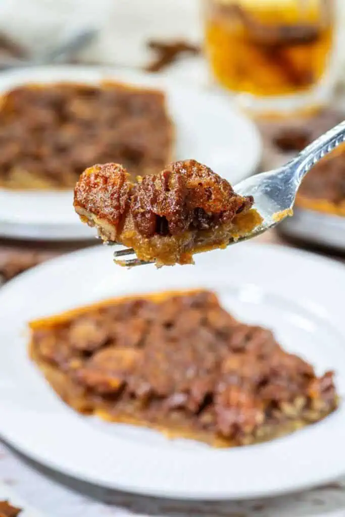 A fork holds a bite of pecan pie above a white plate with a slice of pecan pie; more pie slices are visible in the background.