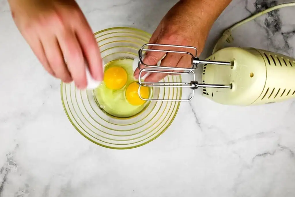 A person cracks eggs into a glass bowl next to an electric hand mixer on a marble countertop.